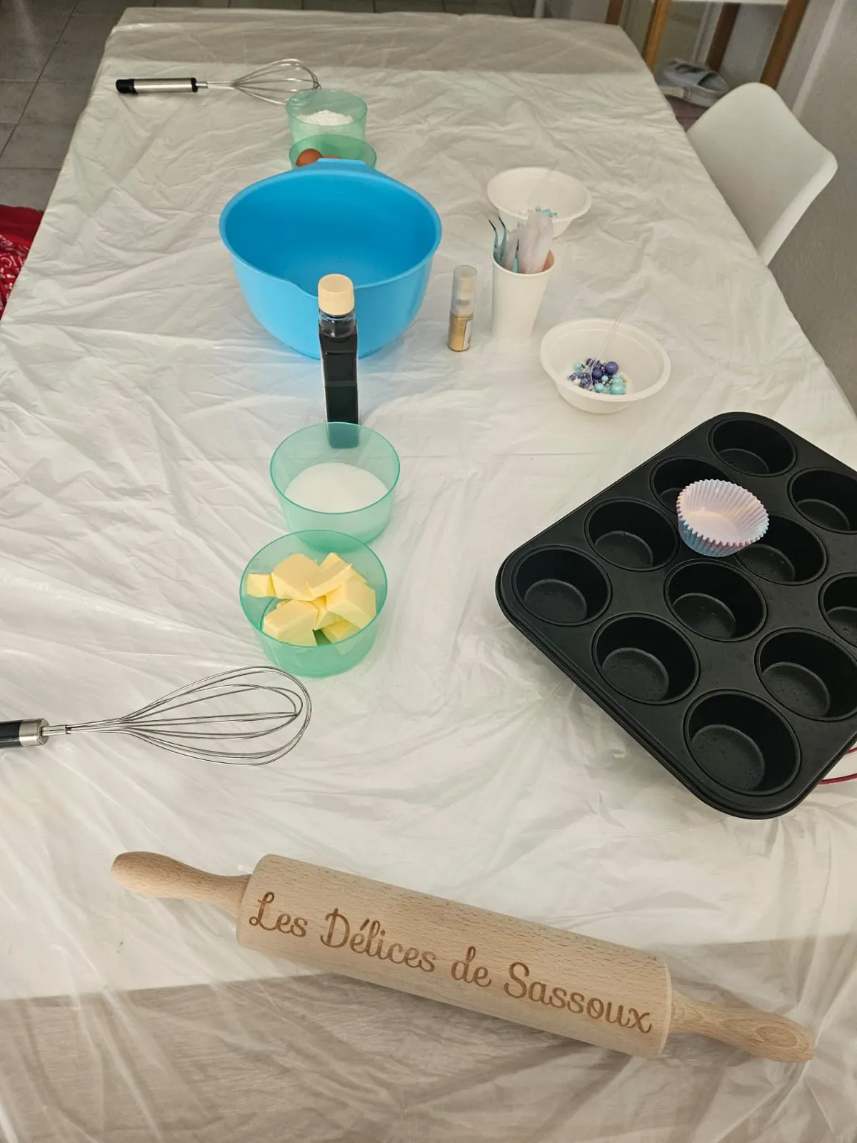 Matériel et ingrédients préparés pour un atelier pâtisserie : bol, fouet, moule à muffins et rouleau personnalisé